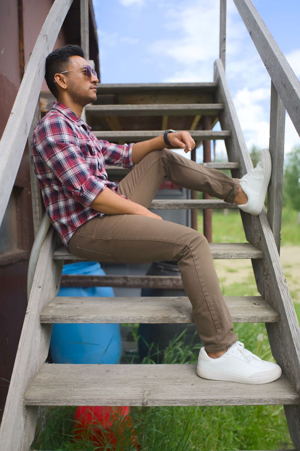 Person in plaid shirt and khaki pants relaxes on wooden steps outdoors with blue barrel in background.