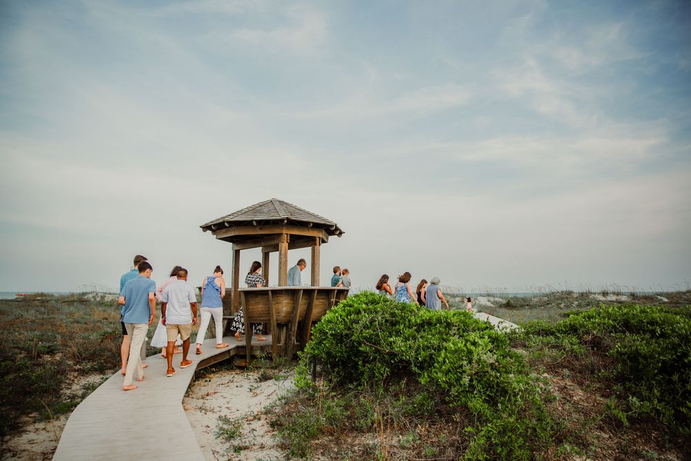 Beachgoers gather at a wooden observation deck overlooking the coastal dunes on a hazy summer day.