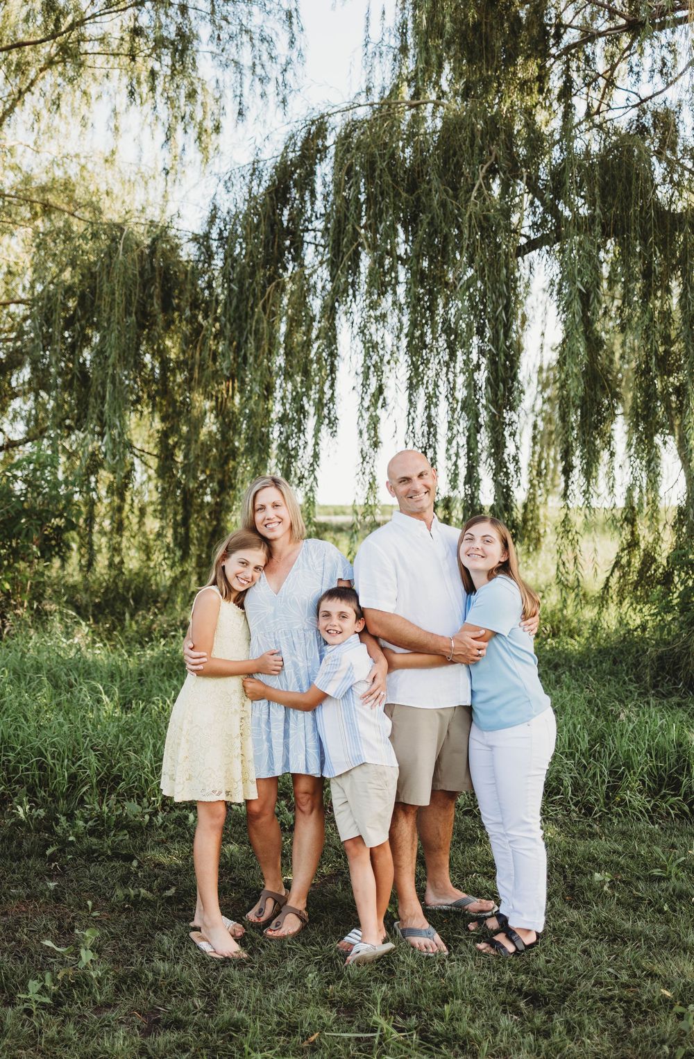 A group in coordinated white and blue outfits poses casually under a weeping willow tree on a grassy hillside.