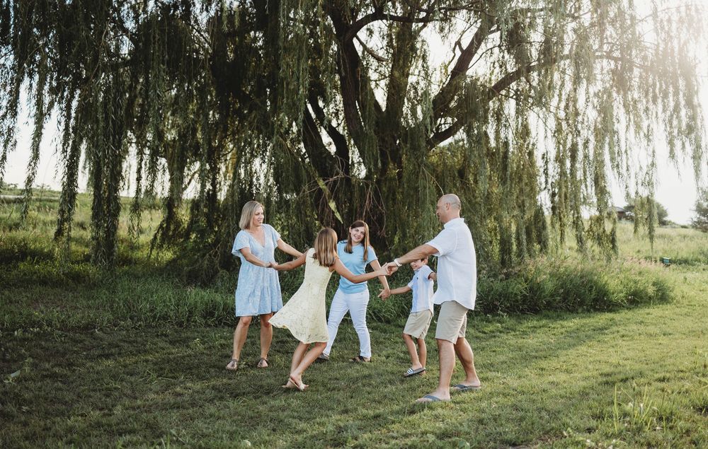 Family plays together under a scenic weeping willow tree in a sunlit meadow on a bright summer day.