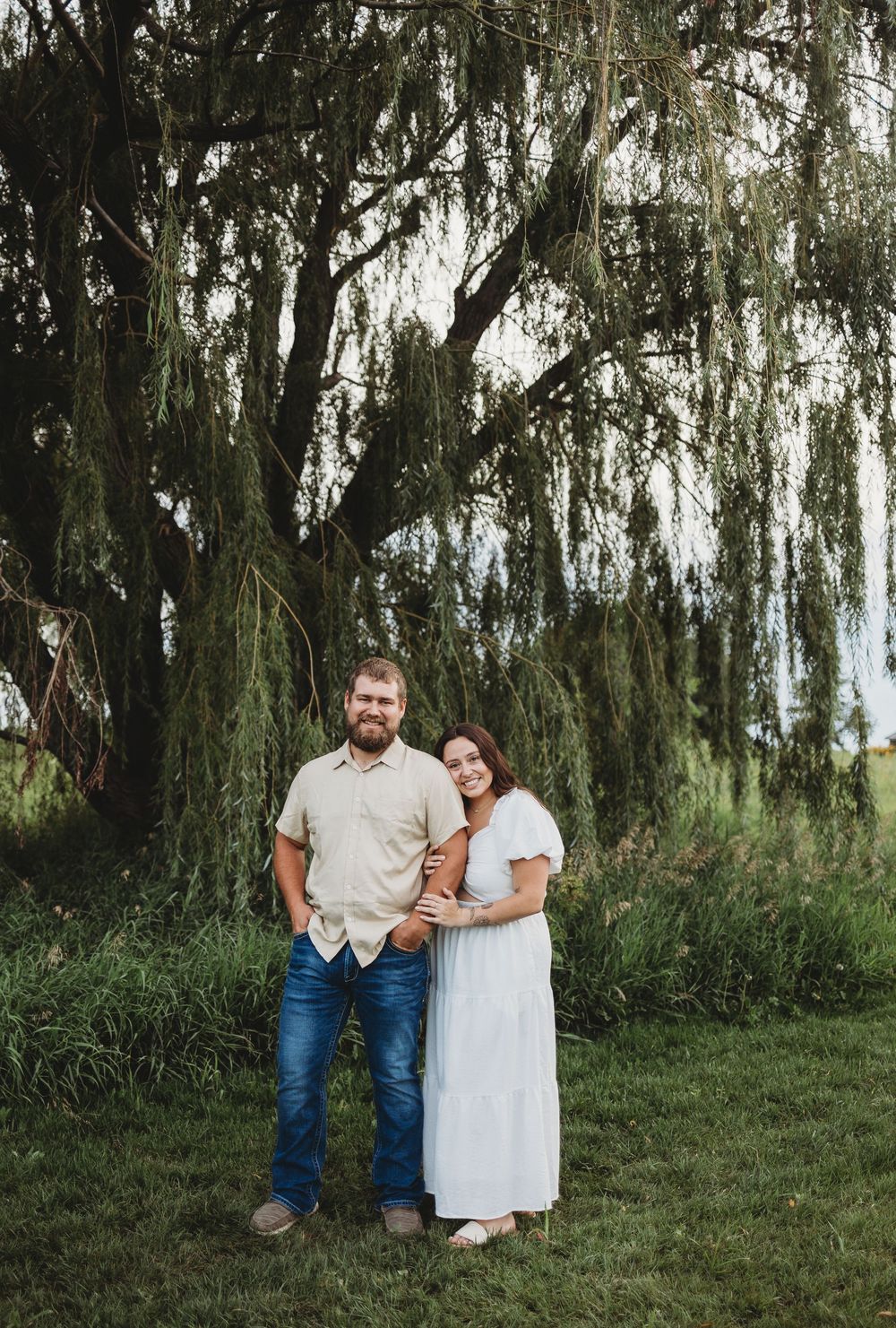 A couple stands together under a large weeping willow tree on a grassy lawn for an outdoor portrait.