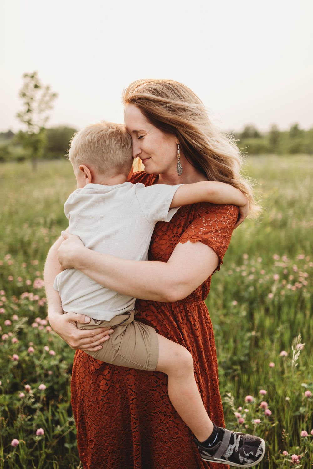 A tender embrace in a wildflower meadow at sunset shows a rust-colored dress against the golden light and blooming field.