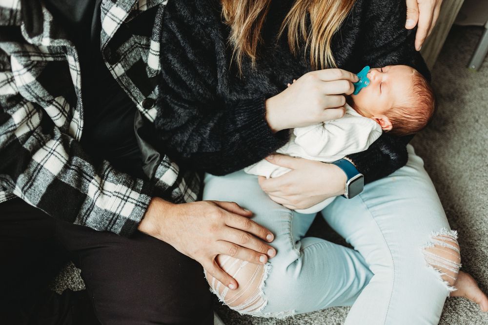 Infant wearing white onesie rests on someone's lap with plaid and black clothing visible.