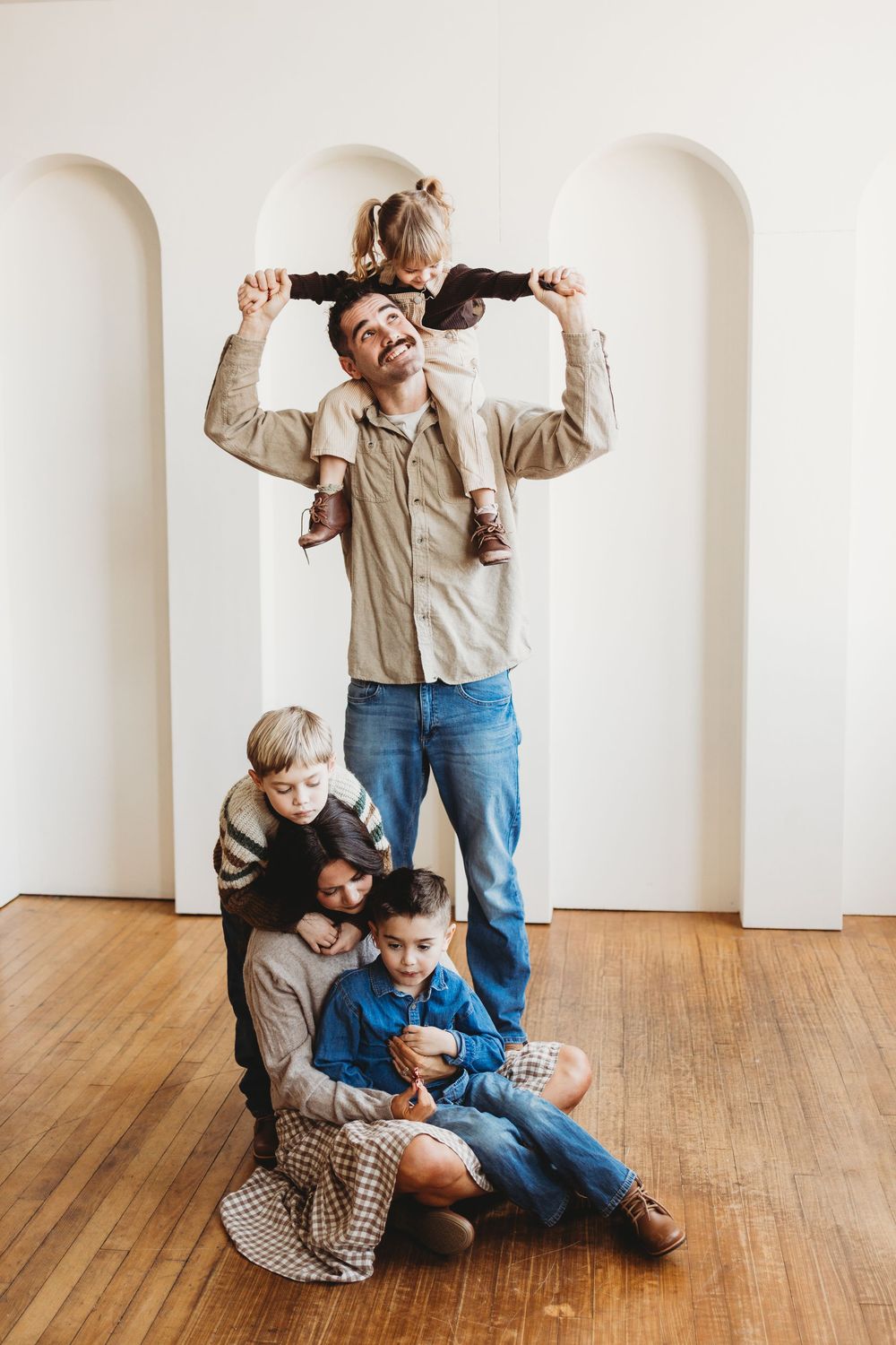 Playful family of four having fun posing and stacking together in front of white archway in minimal interior setting.
