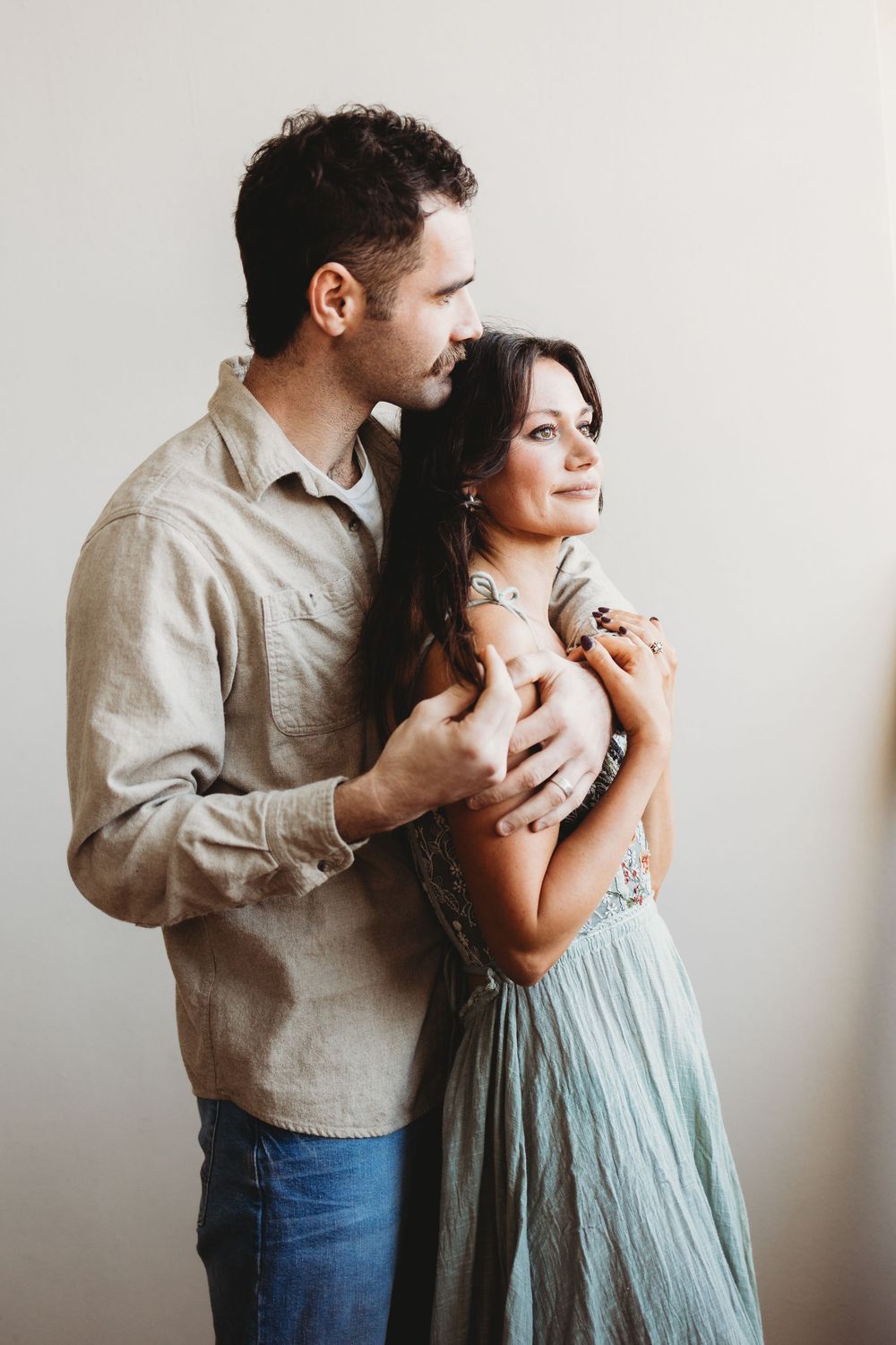 A romantic couple embraces in a minimal studio setting, with her wearing a flowy mint dress and him in casual attire.