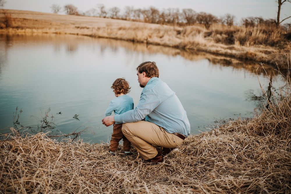 Two people crouched by a calm pond's edge on a golden autumn afternoon, watching ducks swimming in the water.