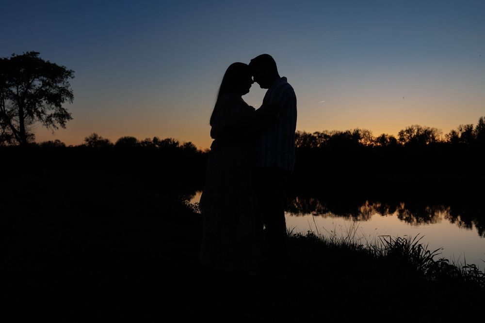 Silhouetted couple embracing by a lake at sunset with trees and reflective water in the background.