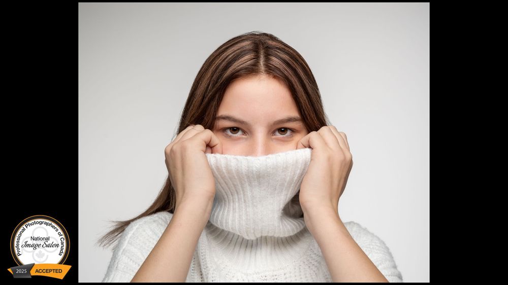 Person in a cozy white turtleneck sweater playfully hiding their smile behind the collar against a light background.