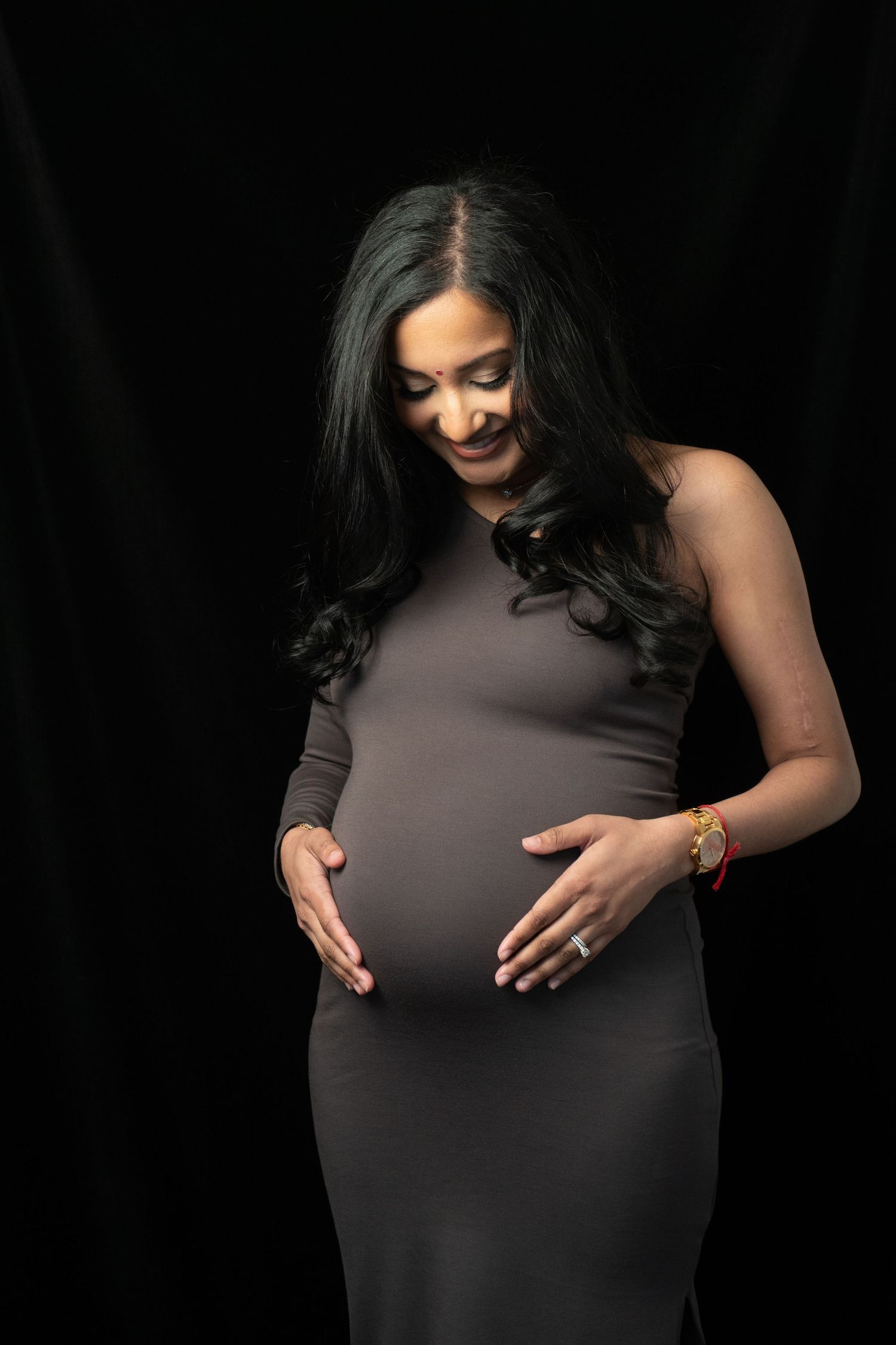 Expectant mother in dark dress touching belly against black background.