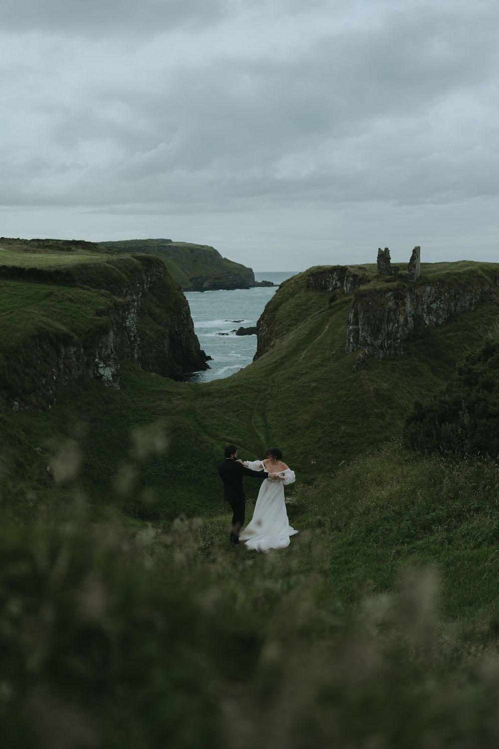 Dramatic coastal cliffs frame two figures in wedding attire overlooking the ocean.