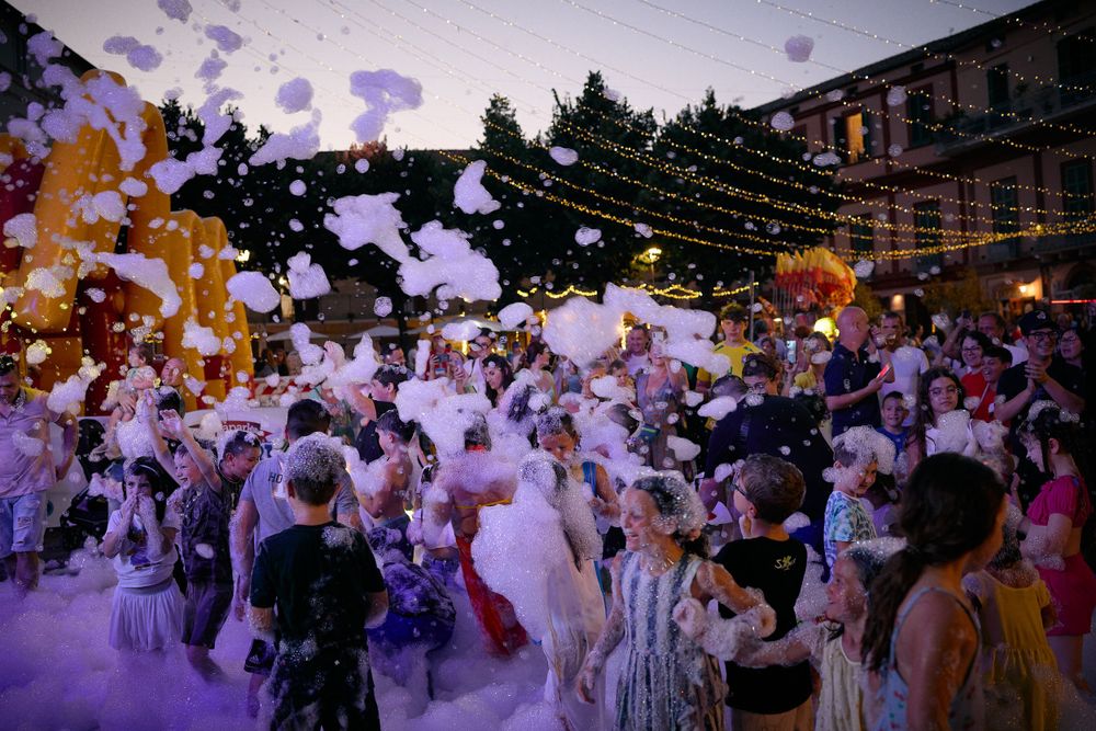 Crowd celebrating at an outdoor party with purple foam and string lights overhead.