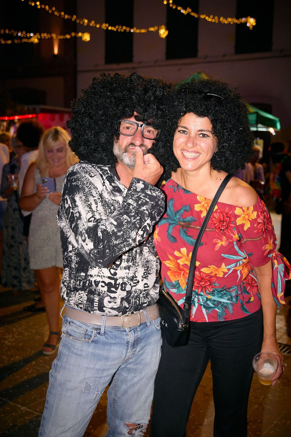 Two partygoers wearing large afro wigs posing beneath string lights at a nighttime event.