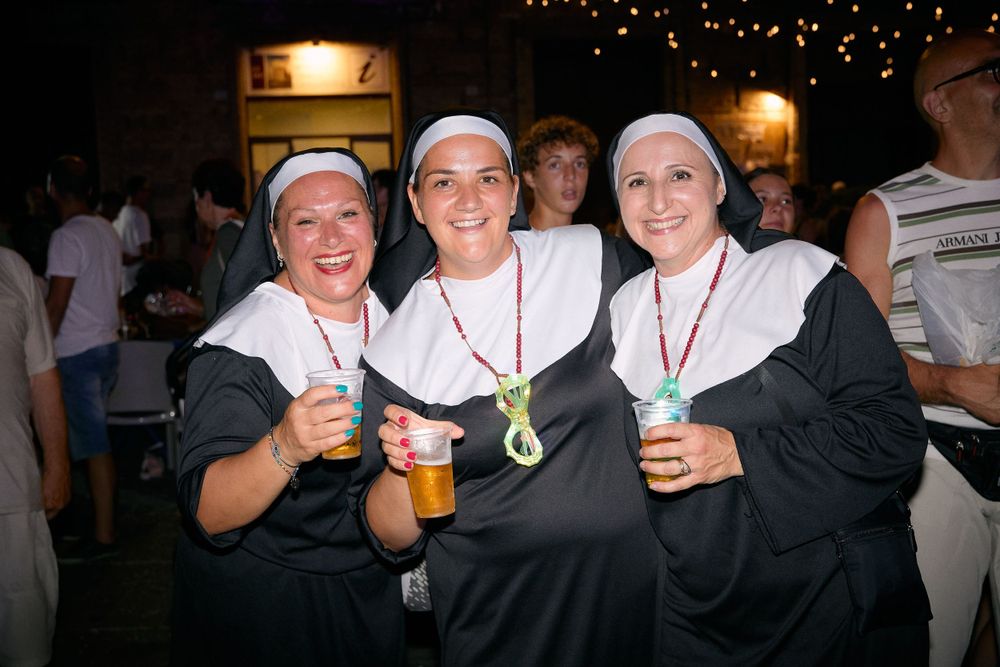 Three people dressed as nuns holding drinks and posing at a party under string lights.