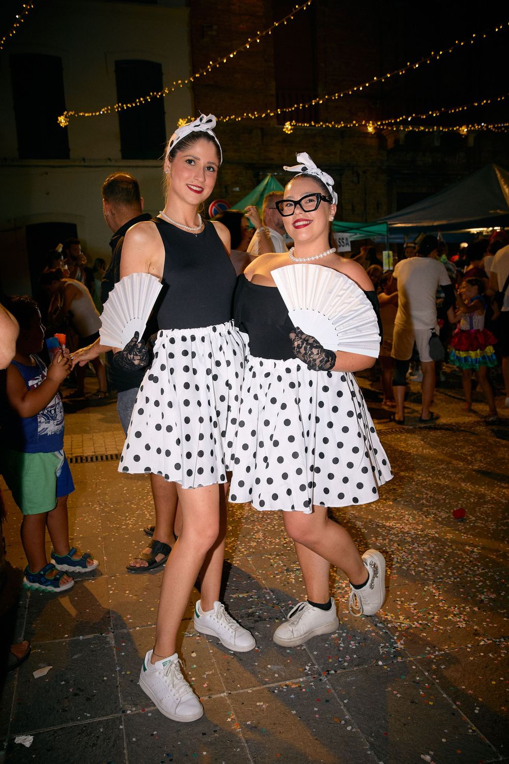 Two people in matching polka dot skirts and white accessories pose under string lights.
