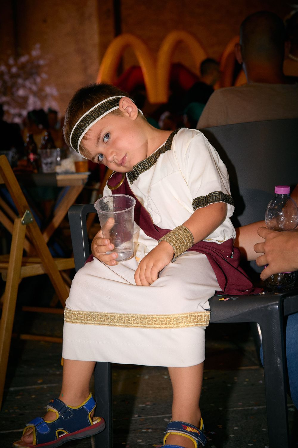 Child in a Greek-inspired costume sitting on chair at event with McDonald's sign visible.
