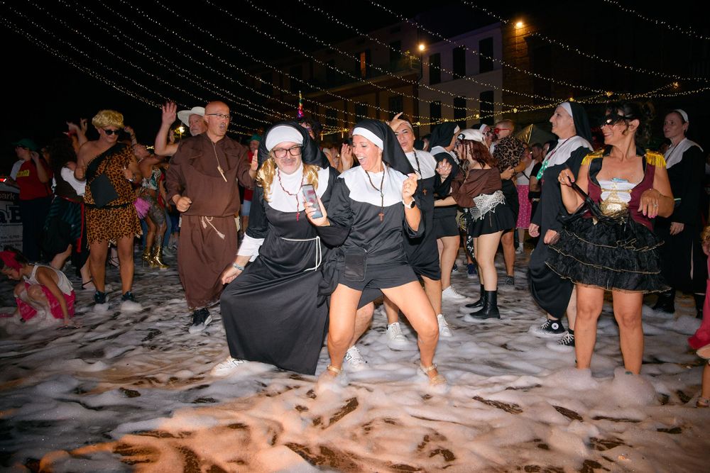 A lively nighttime dance party with people in costumes dancing on sandy ground under string lights.