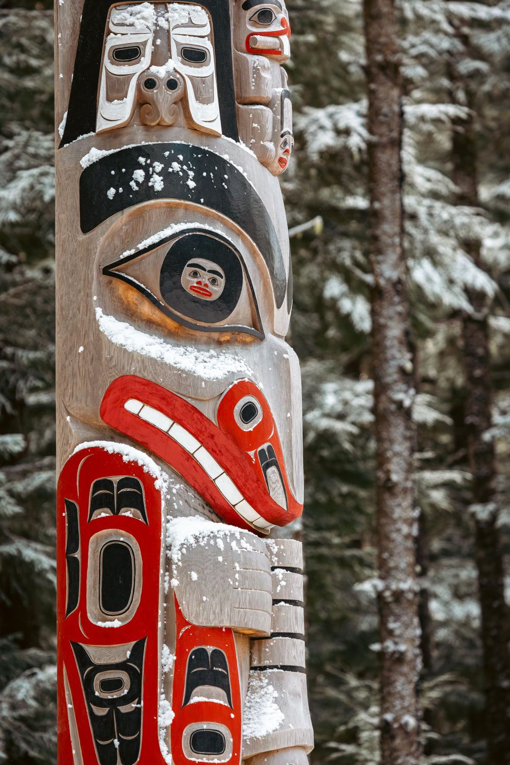 Colorful traditional Pacific Northwest totem pole with red and black designs dusted with snow in a forest setting.