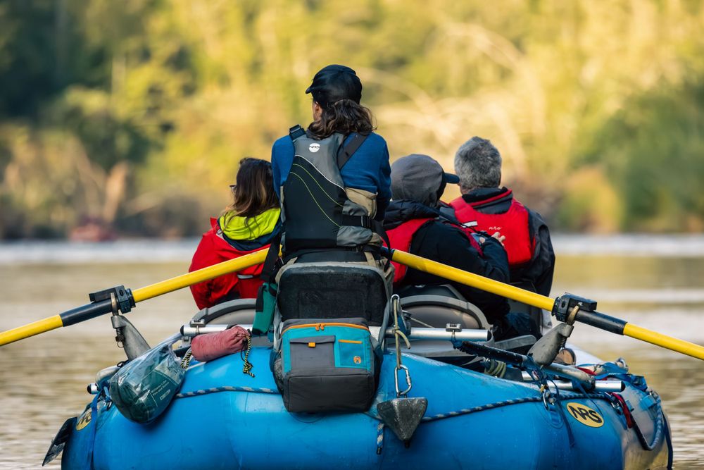 People rowing and navigating a blue raft boat with gear and equipment loaded on the Atnarko river in Bella Coola.