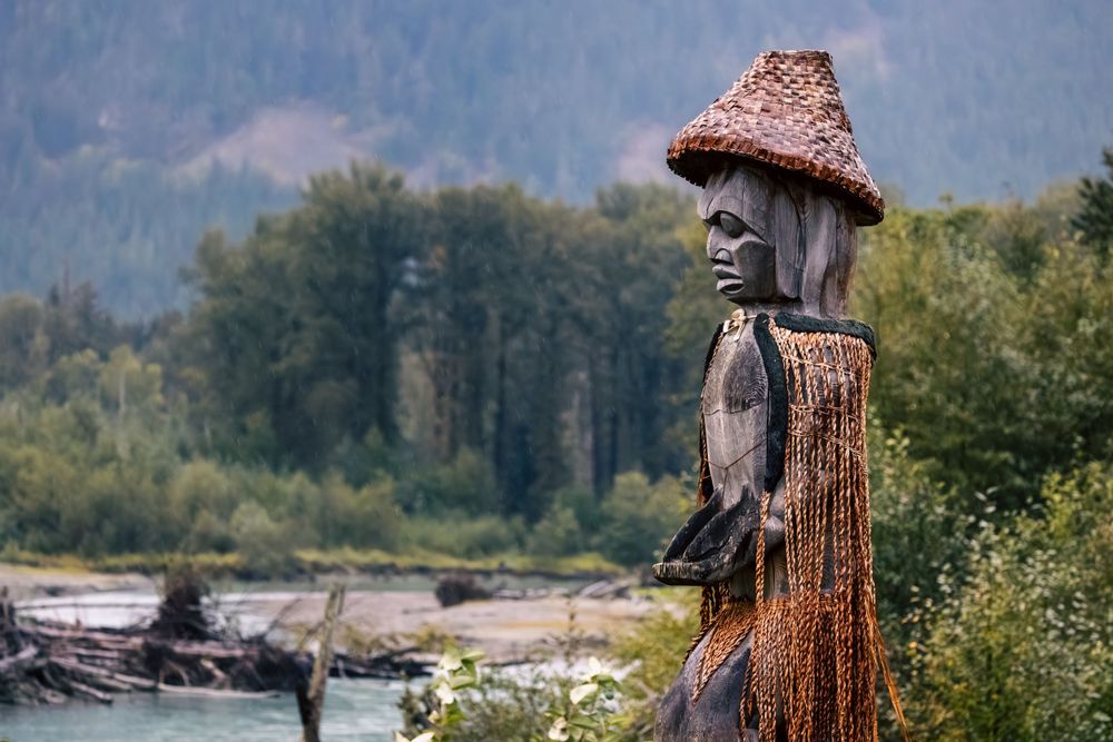 A wooden totem pole with a woven hat stands near a river with mountains and forest in the background in Bella Coola.