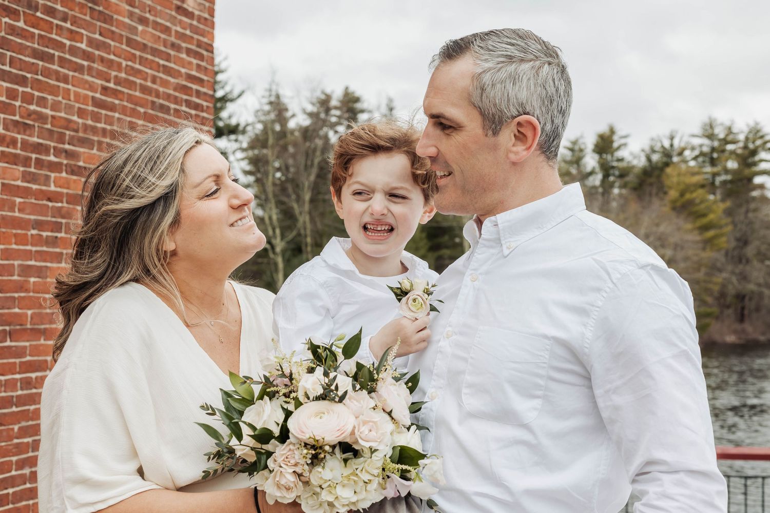 A joyful winter wedding moment showing a couple and child laughing together with a white floral bouquet against brick wall.