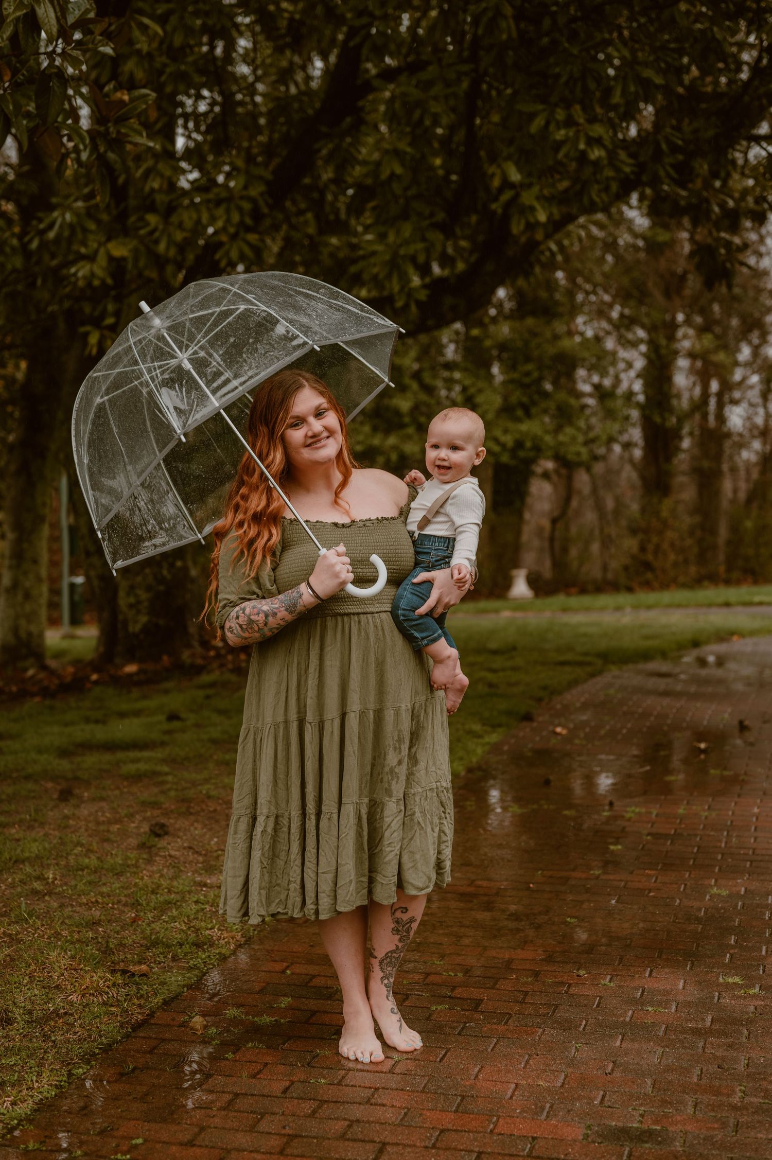 A woman in a green dress holds a clear umbrella while standing barefoot on a brick path with a baby. photo by Samantha Campbell imagery