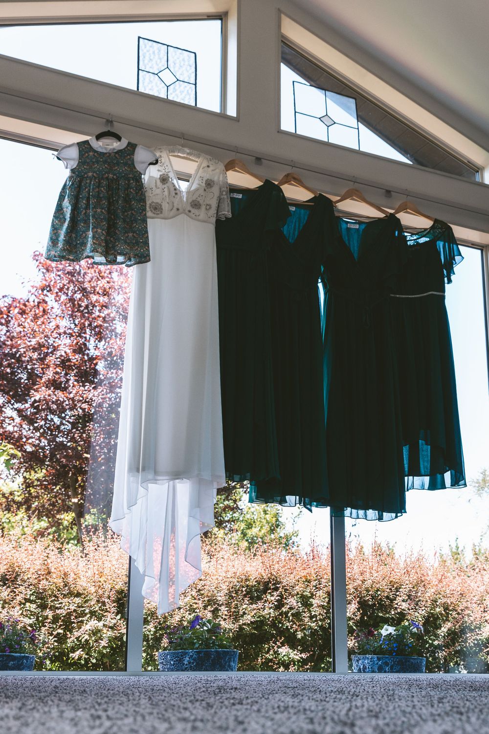 Green flower girl dress and dark bridesmaid dresses displayed against bright window.