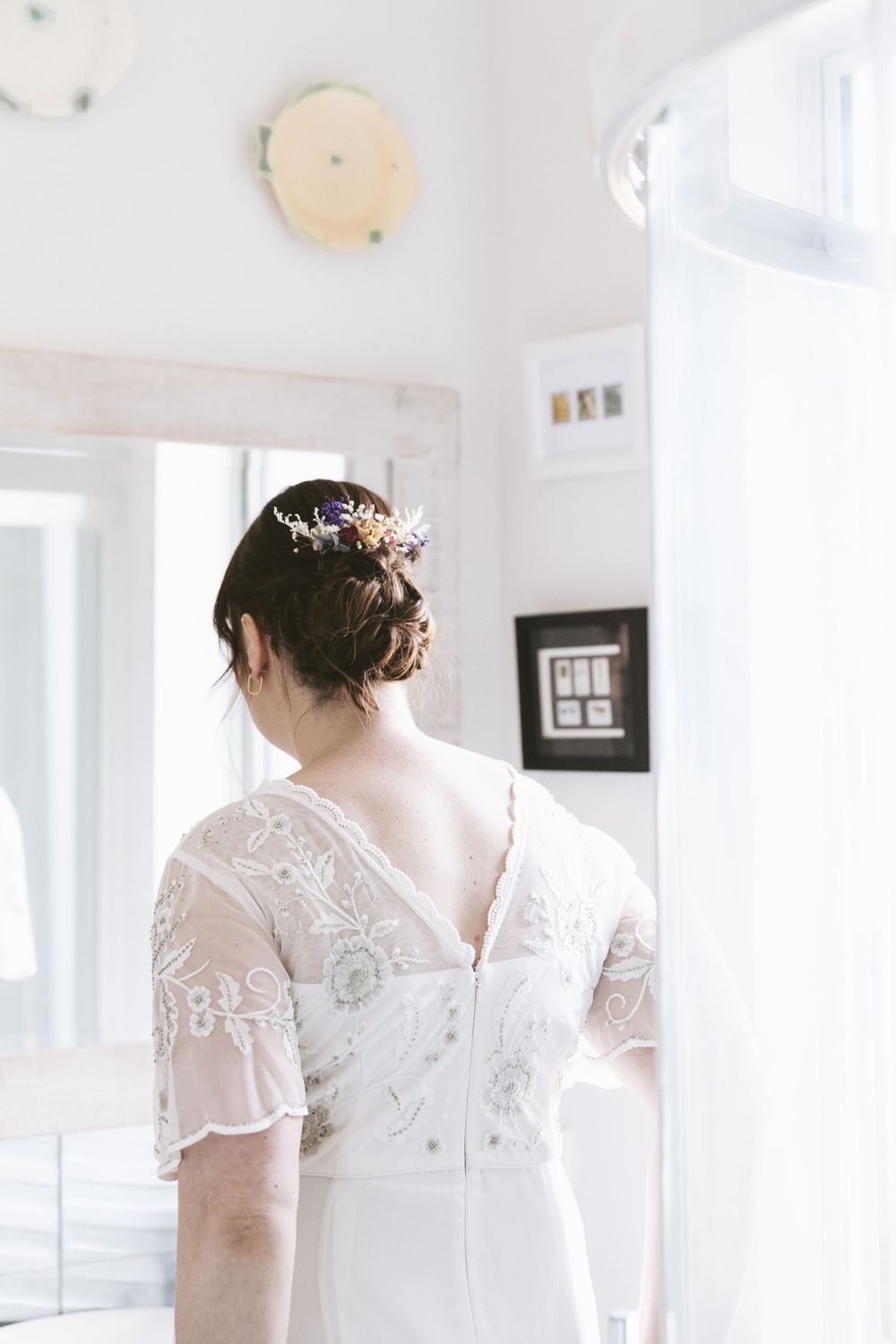 Back view of a bride wearing a delicate lace wedding dress with floral hair accessory in a bright room.