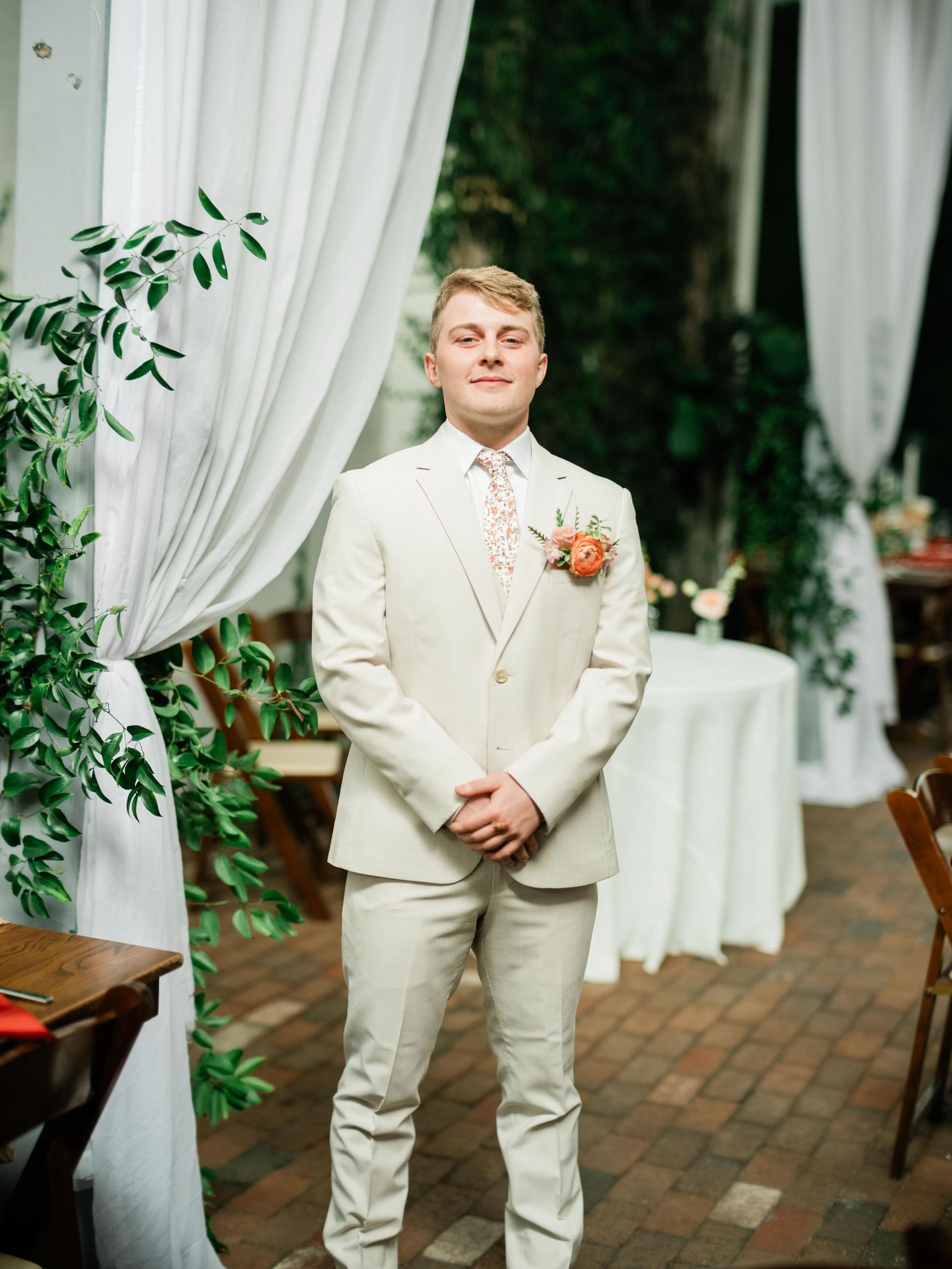 Groom in cream colored suit stands in front of white curtains and greenery at wedding venue.