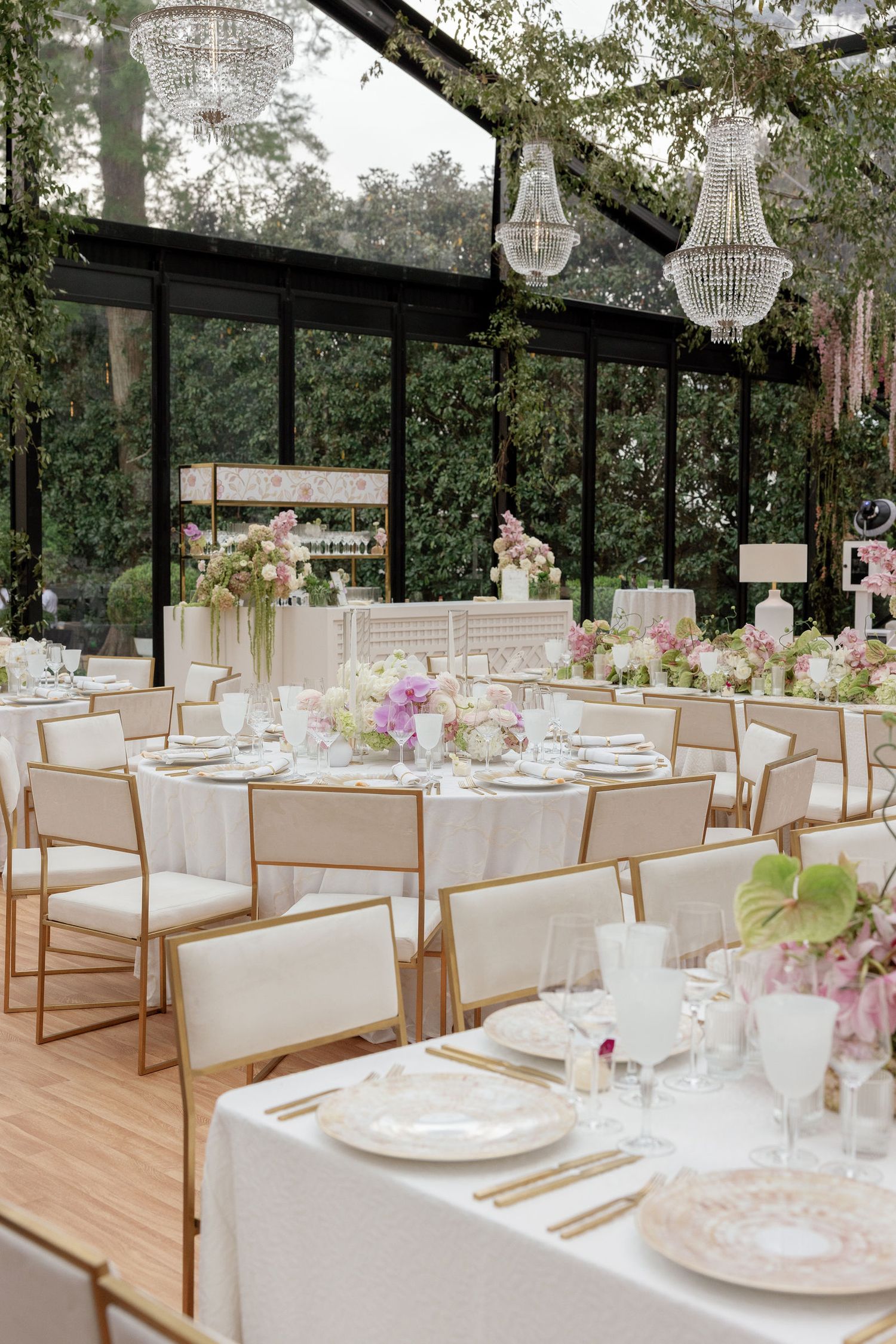 Round dining tables with white linens and gold chairs set for an elegant garden reception beneath hanging crystal chandeliers.