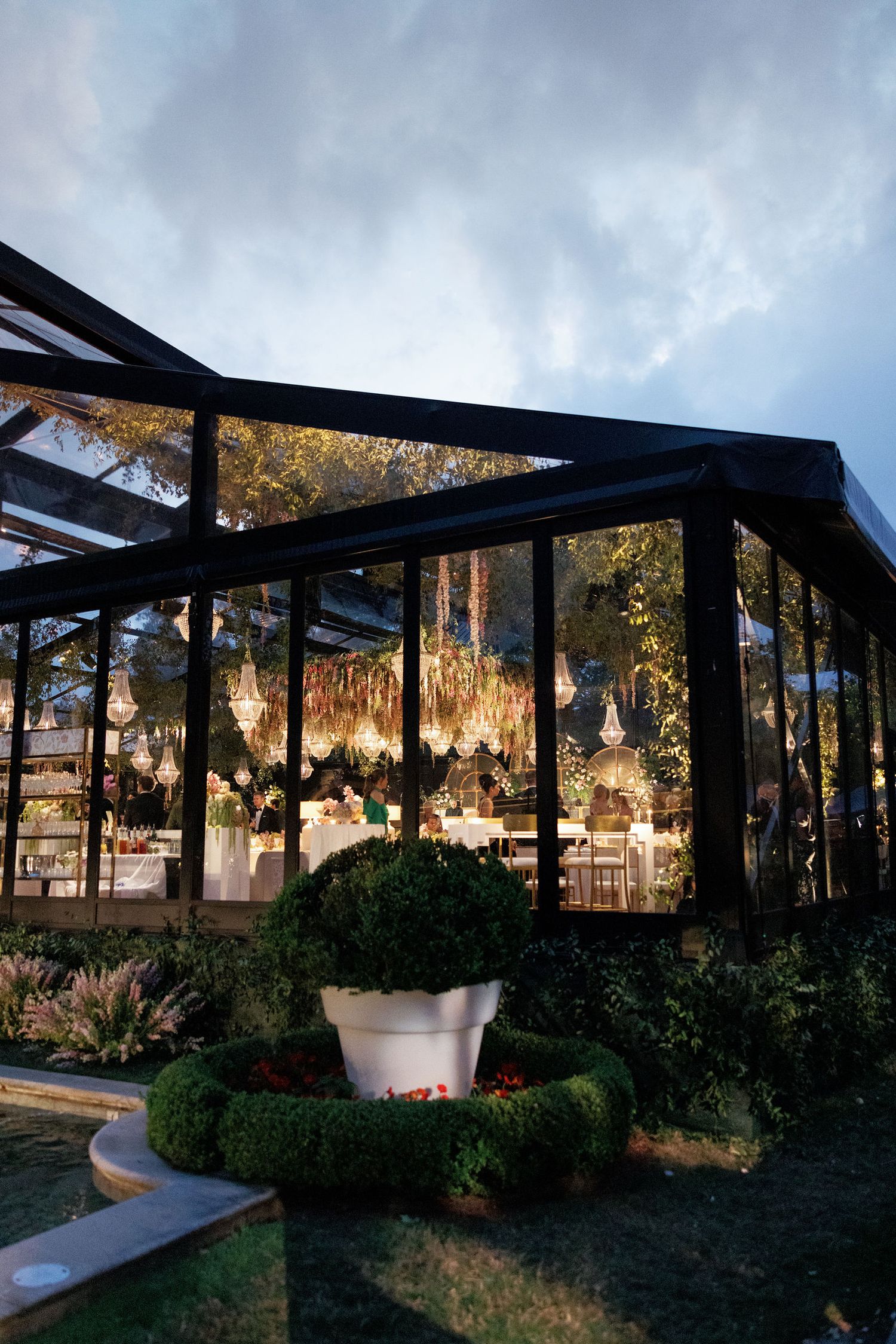 Modern outdoor dining pavilion with black steel frame and glass panels surrounded by lush landscaping at dusk.