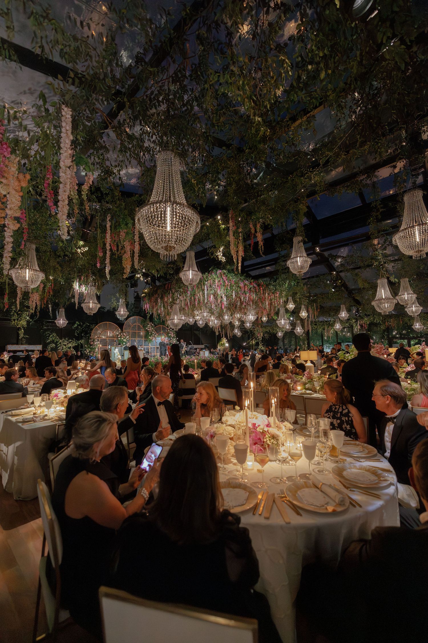 Elegant indoor wedding reception with crystal chandeliers and greenery hanging from the ceiling over round dining tables.