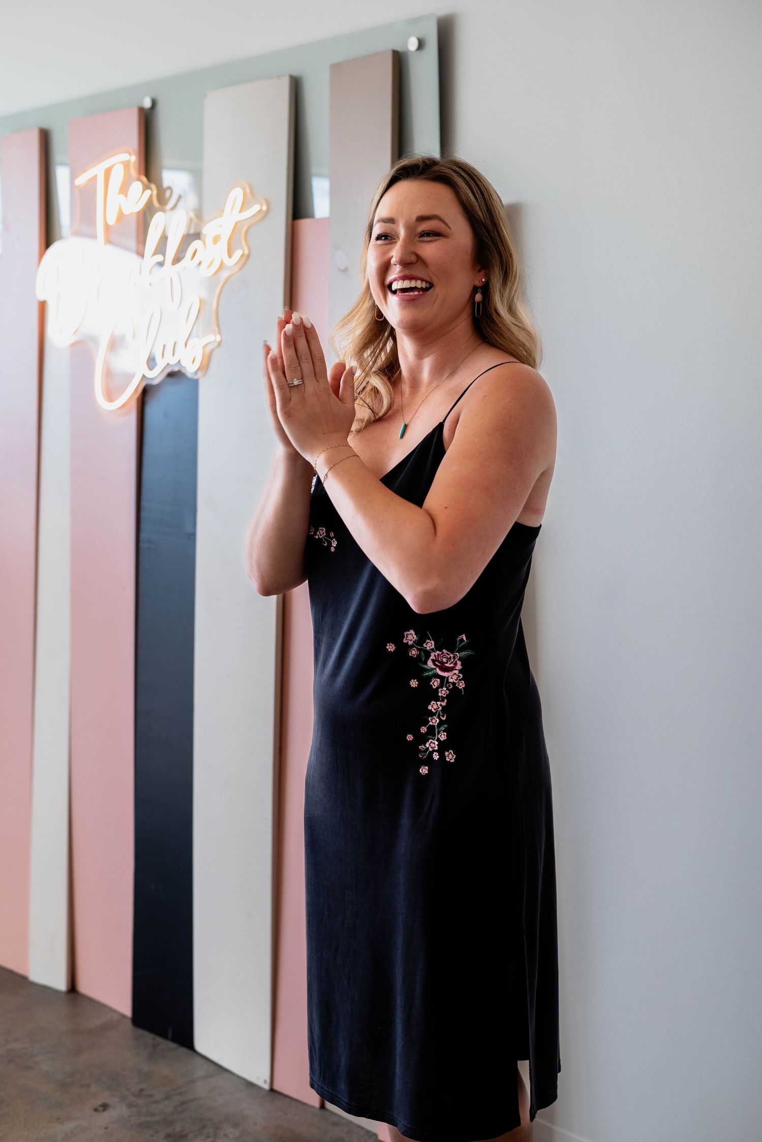 Person in black dress laughing joyfully against pink and white striped wall with neon sign.