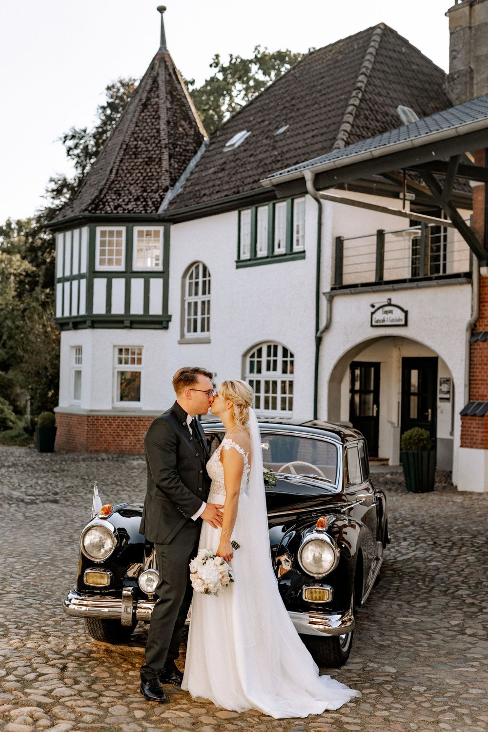 Couple poses with vintage black car in front of Tudor-style wedding venue.