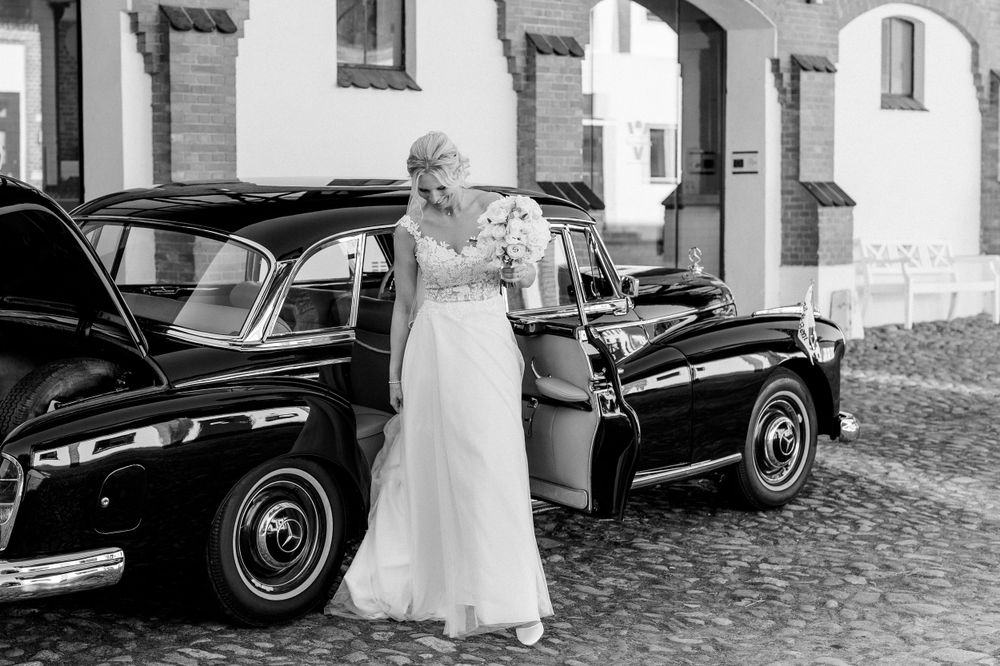 Elegant bride in lace gown poses beside classic vintage black car.