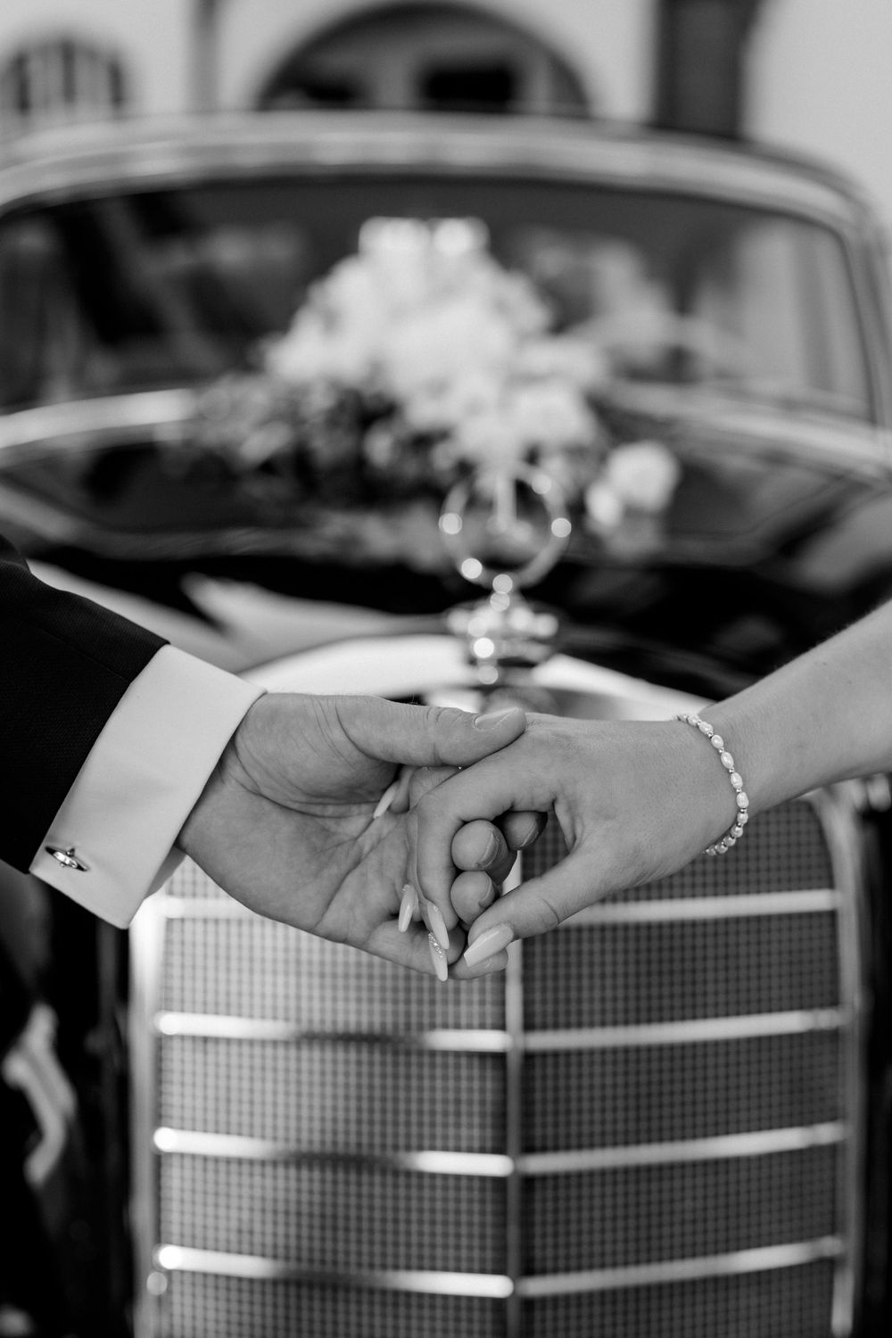 Close-up of wedding couple holding hands in front of vintage car grille.