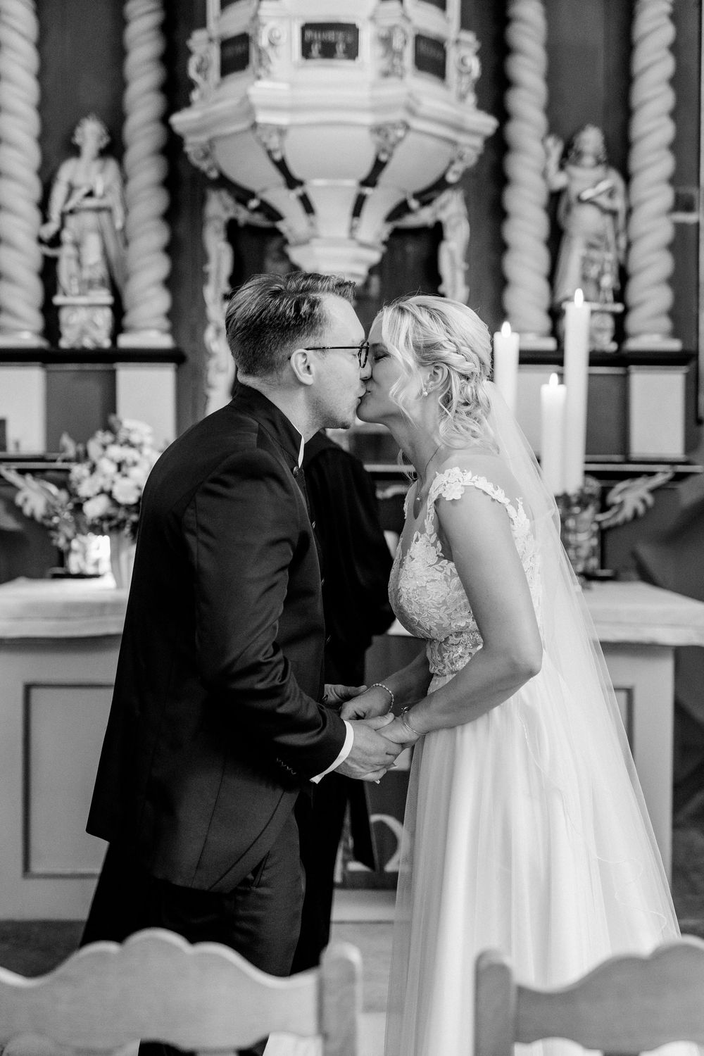 A tender black and white wedding ceremony photo captured inside a church with ornate decor and statues in the background.