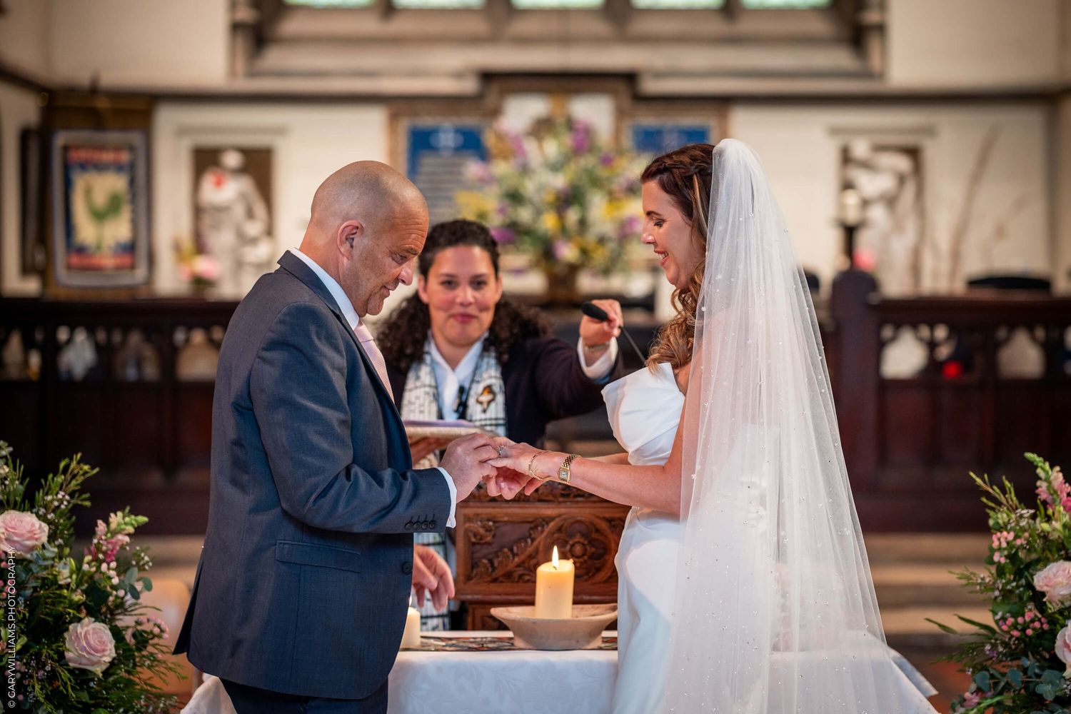 A couple exchanges rings during their wedding ceremony inside a church decorated with floral arrangements.