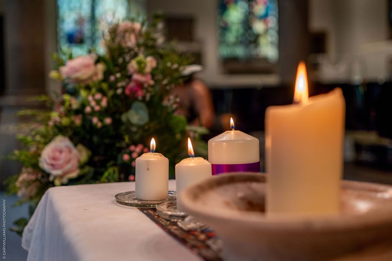Lit white candles and pink floral arrangements on white tablecloth in church setting with stained glass windows in background.