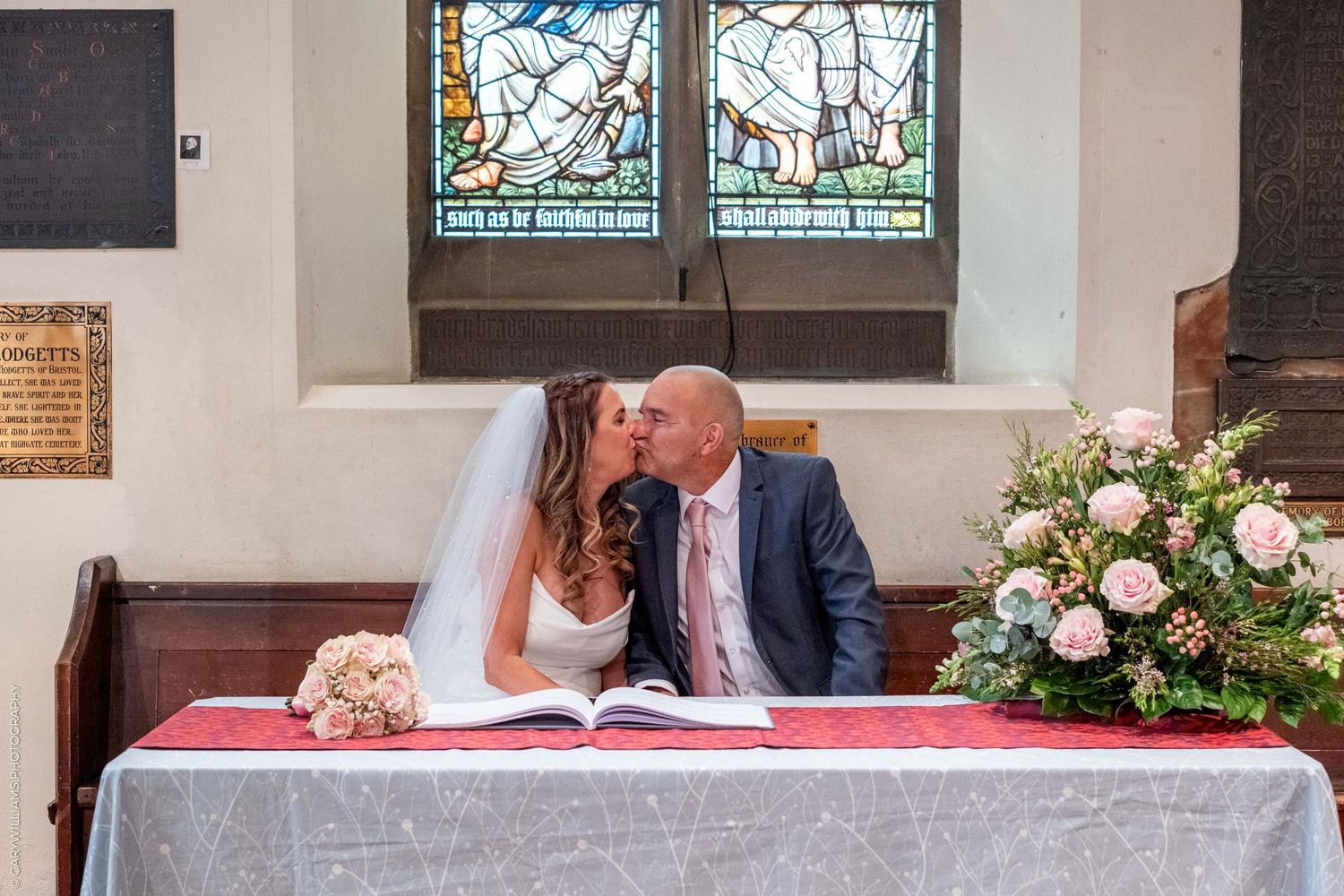 Two people sit together at an altar with floral arrangements and stained glass windows in a beautiful church setting.