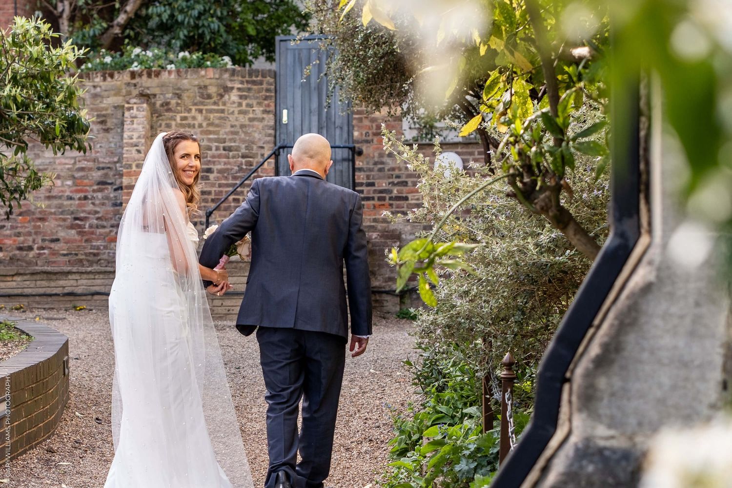 A bride in a white lace gown walks up garden steps with a gentleman in a navy suit on a sunny day.