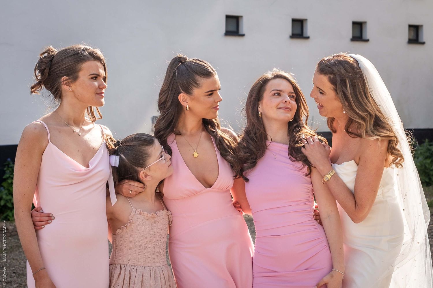Group of people in pink and blush-colored dresses standing together and smiling while gathering outdoors on sunny day.
