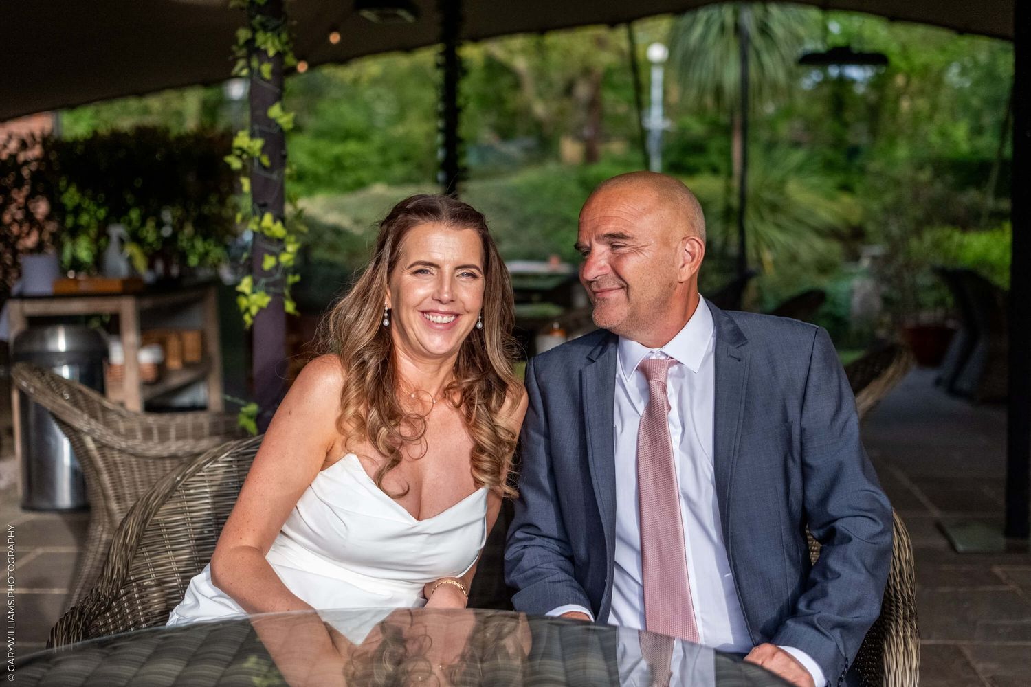 A couple in wedding attire share a joyful moment while sitting together at an outdoor covered patio.