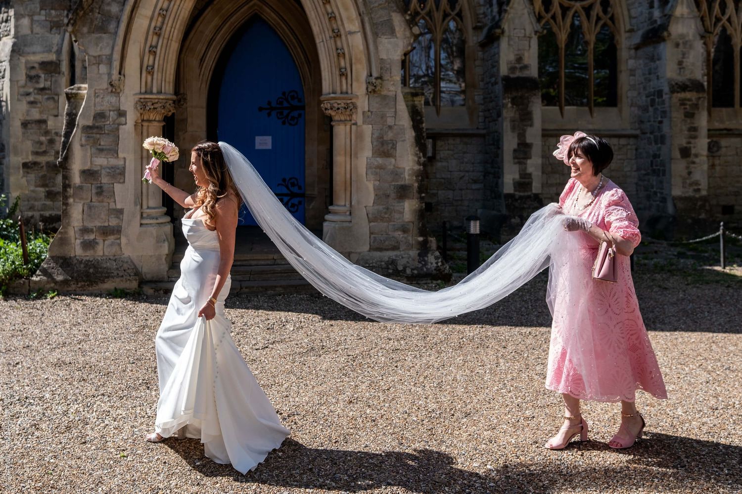 Someone wearing a flowing white wedding dress stands with a person in pink holding their long veil outside a gothic church.