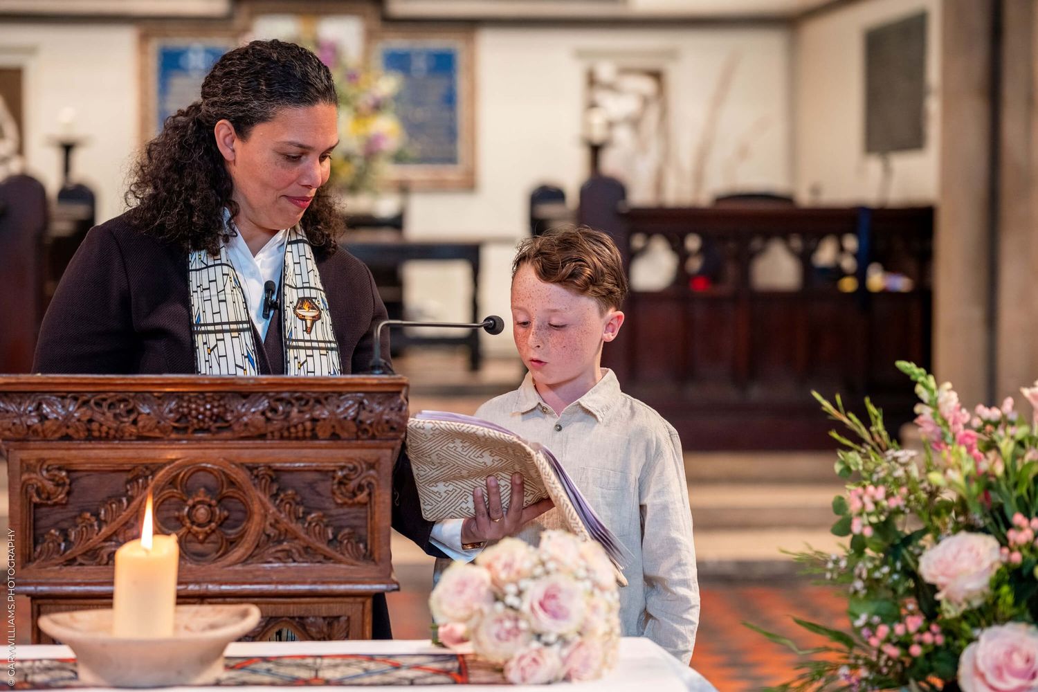 Two people stand at an ornately carved wooden pulpit with floral decorations in a church setting.