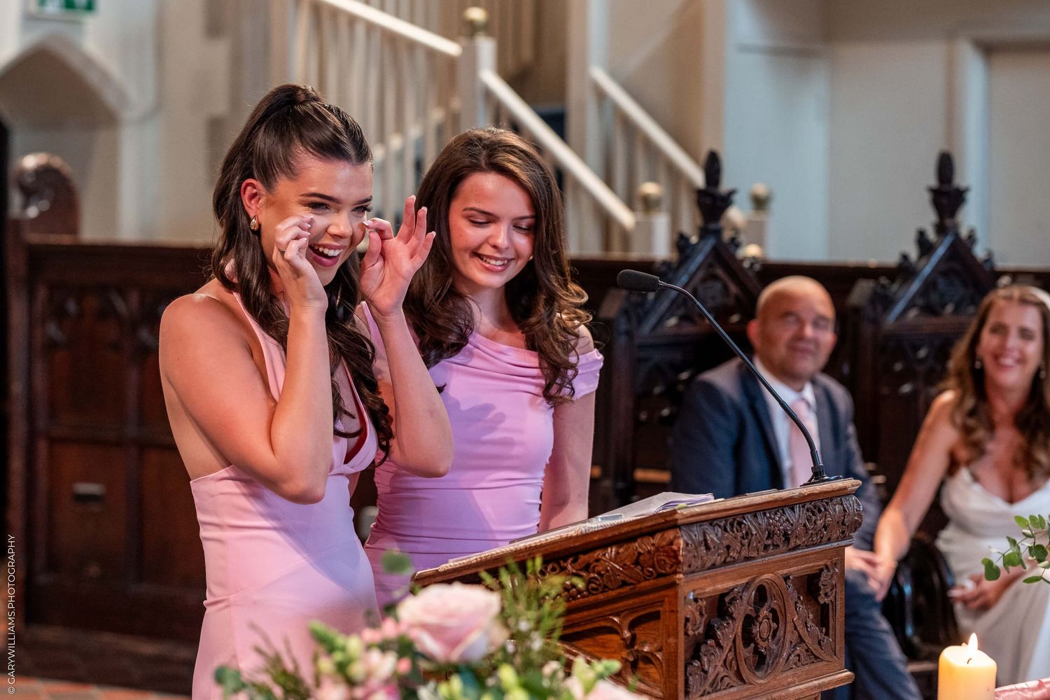 Two attendees in pink dresses share an emotional moment during a wedding ceremony in an ornate church.