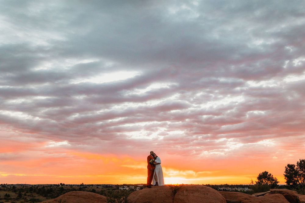 A silhouetted couple embraces on a hill against a dramatic orange and pink sunset sky with wispy clouds.