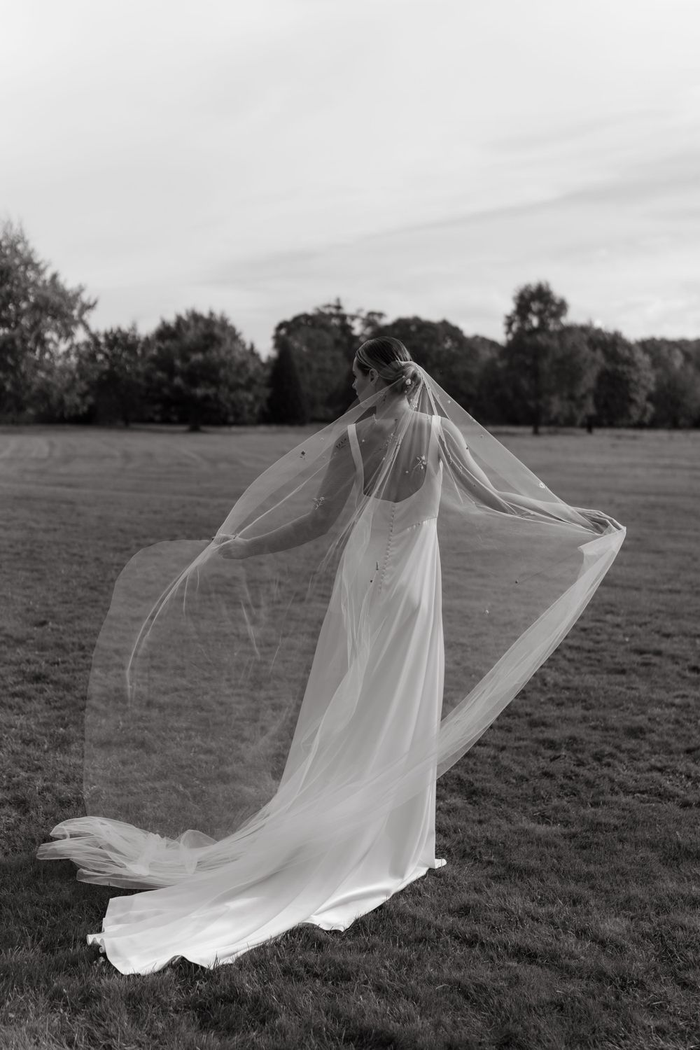 A flowing bridal veil catches the wind as a bride stands in a grassy field in black and white.