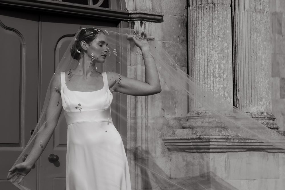 A bride in a sleek white wedding dress poses elegantly against a classical stone building exterior.