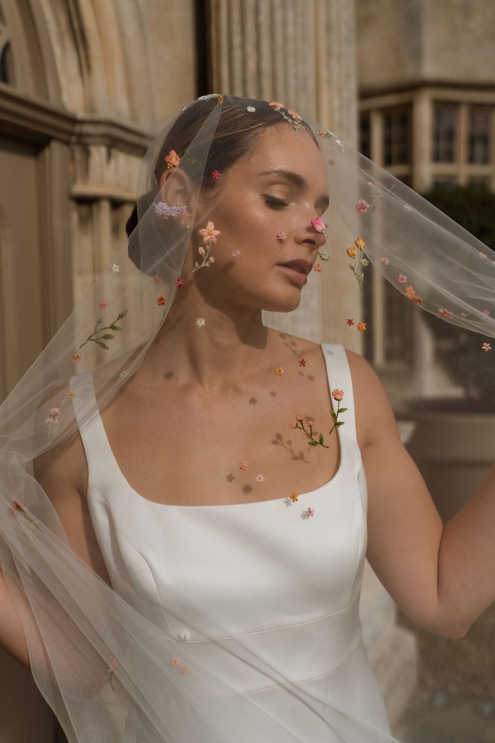 Elegant bridal portrait featuring white square-neck gown and delicate veil with golden floral details in soft lighting.