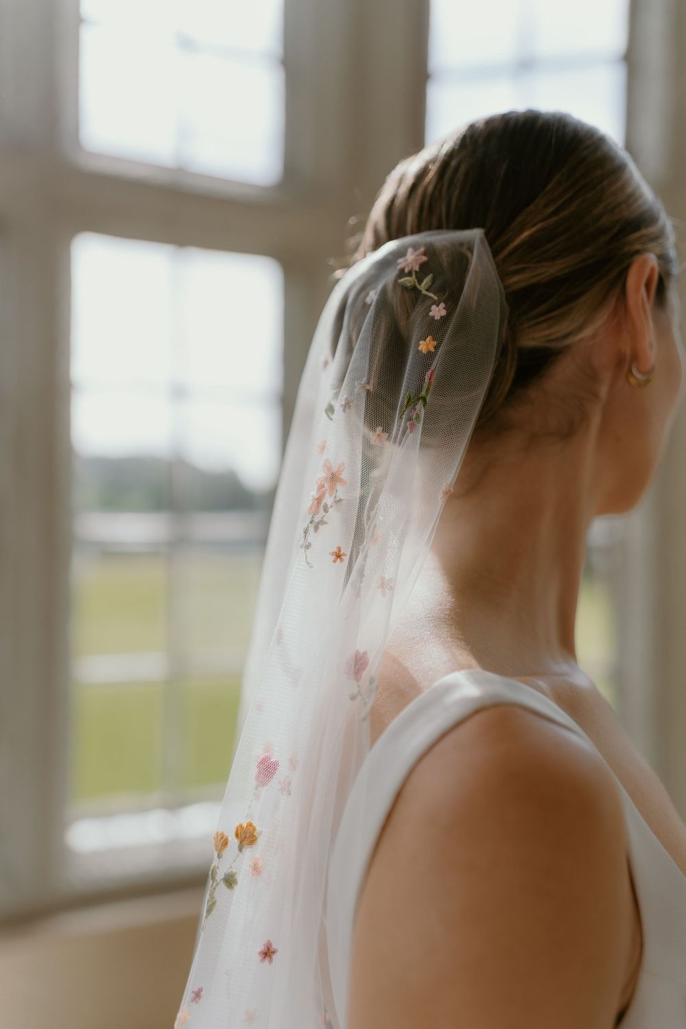 A bride wearing a delicate floral embroidered veil stands by a window in soft natural light.